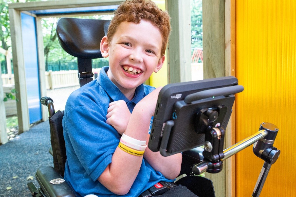 Charlie uses a Talk Pad mounted to his wheelchair for easier access to Grid for communication. You can see the blue accessibility buttons. Charlie is a young boy with his blue school jumper on, he is smiling.