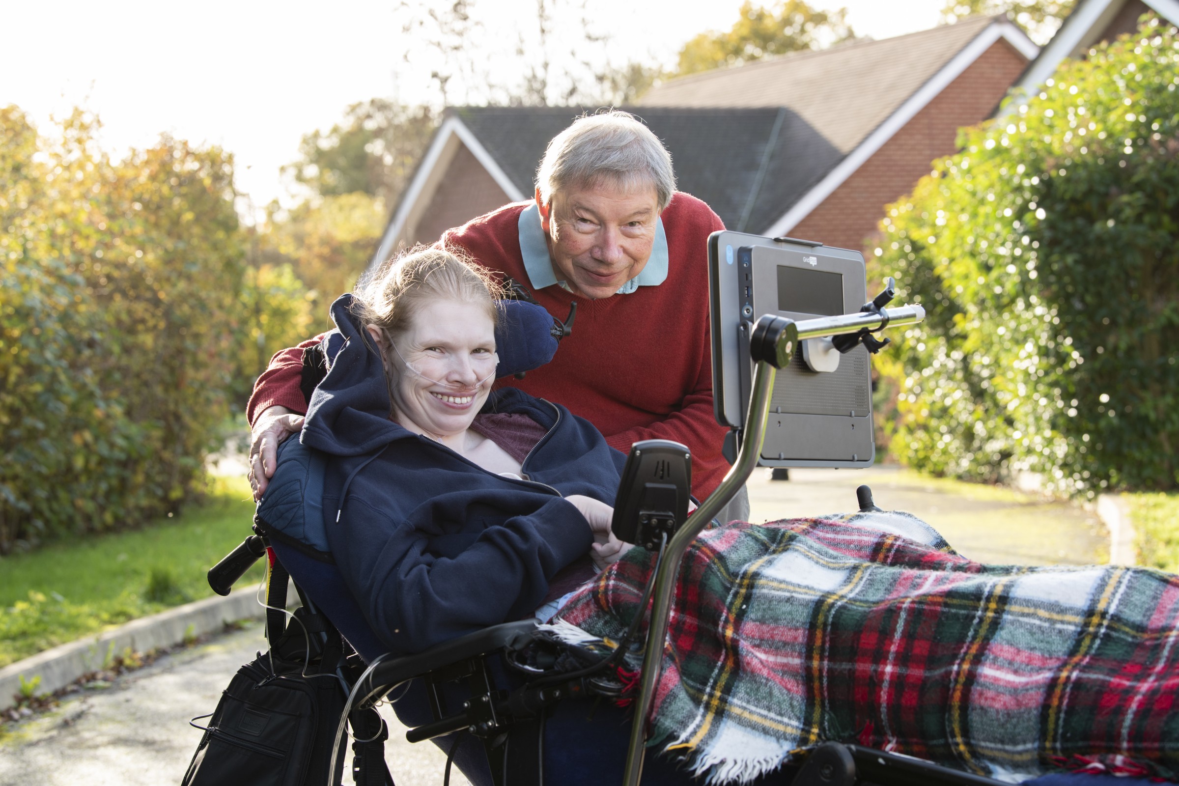 Two people smiling in a leafy residential area. The woman in the foreground is in a wheelchair with a Grid Pad 13 mounted to it.