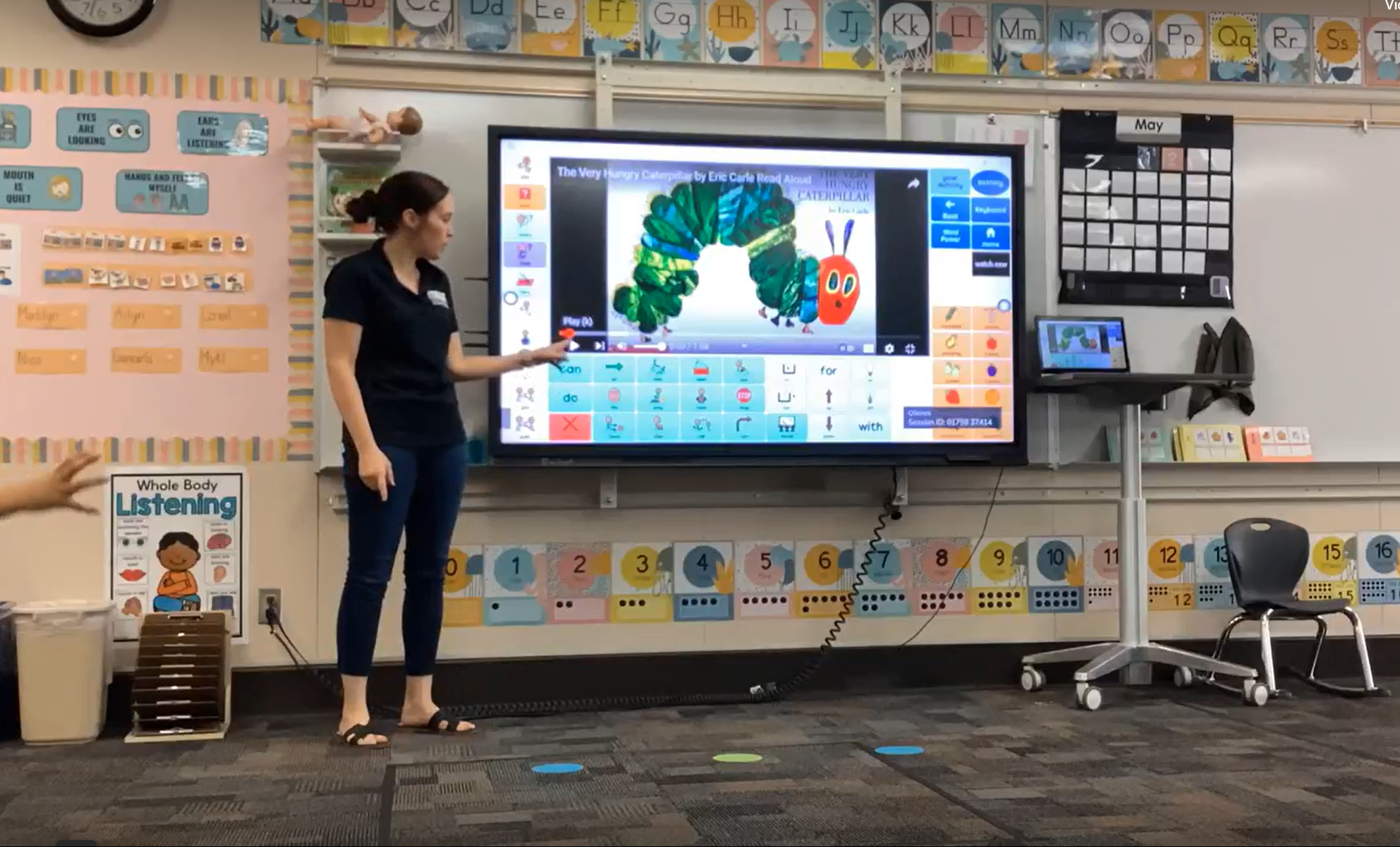 Teacher at the front of a colorful classroom, with the Very Hungry Caterpillar shown in Grid software on an interactive whiteboard. A video of the book is in the middle of the screen, with relevant words and phrases surrounding it.