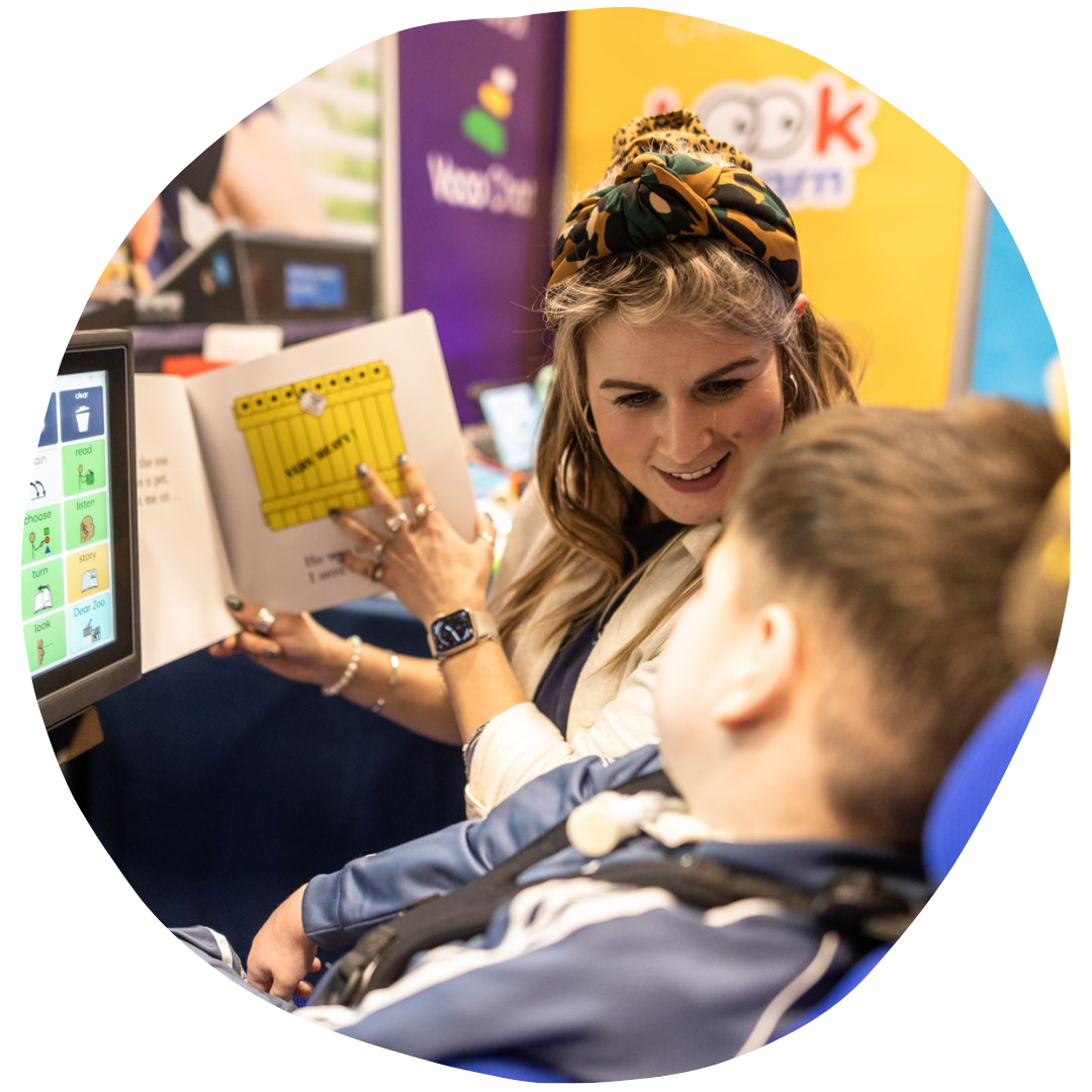 Woman holding up a book next to an AAC device, AAC user is looking at the book