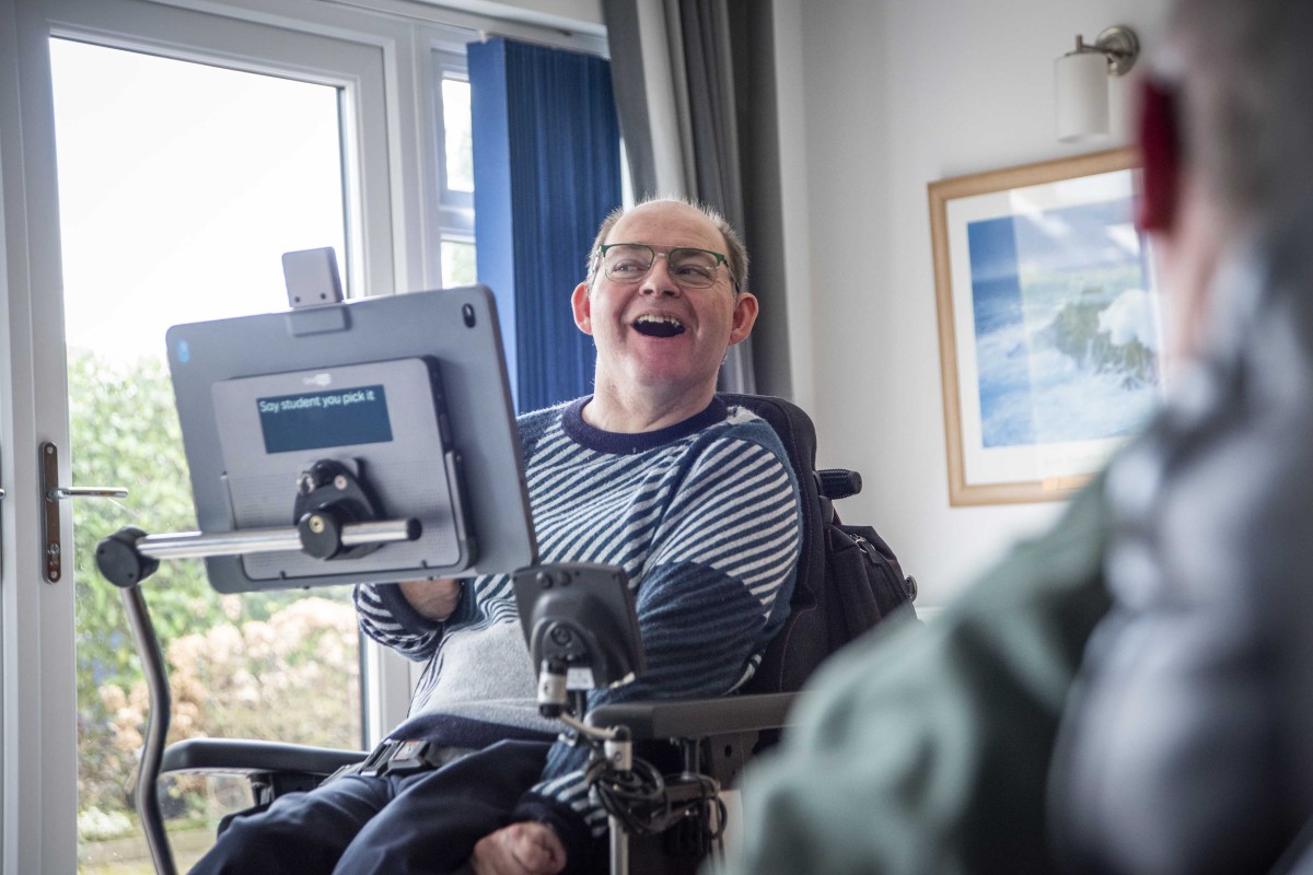 Richard, 44, communicating with someone using his Smartbox Grid Pad mounted to his chair, looking very happy.