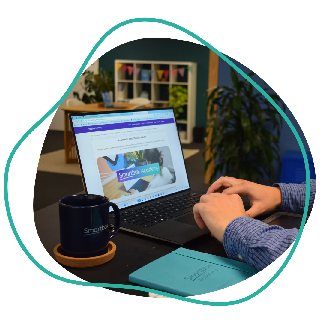 Laptop with Smartbox Academy on the screen, two hands sitting on top of the keyboard. A Smartbox mug and a notebook are also on the black table.